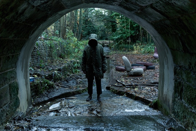 Osoba w płaszczu z kapturem stoi pod tunelem w zalesionym terenie, odwrócona tyłem. Woda płynie pod ich stopami, a porzucone krzesła są porozrzucane na zewnątrz w zarośniętym, ponurym otoczeniu.