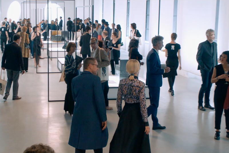Duża grupa ludzi w formalnych strojach przechadza się i rozmawia w jasnej, nowoczesnej galerii sztuki z minimalistycznymi wystawami i wysokimi oknami.