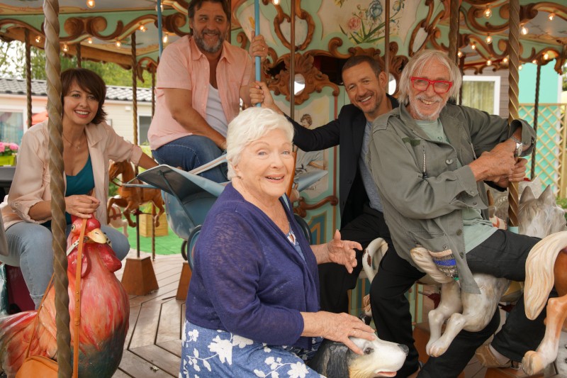 Pięciu uśmiechniętych dorosłych, w tym trzech mężczyzn i dwie kobiety, jedzie na kolorowych figurach zwierząt na zabytkowej karuzeli. Wyglądają na radosnych, cieszących się zabawnym otoczeniem. W tle widać część ozdobnych dekoracji karuzeli.