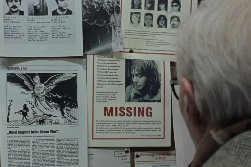 Osoba o siwych włosach patrzy na tablicę ogłoszeń pokrytą czarno-białymi wycinkami z gazet i dużym plakatem informującym o zaginięciu osoby, na którym widnieje zdjęcie młodej kobiety.