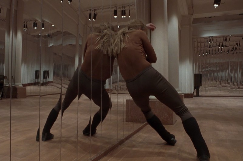 Osoba ubrana w brązowo-czarne ubranie opiera się o lustrzaną ścianę w dużym, pustym pokoju, tworząc na lustrach liczne odbicia swojej pozy.