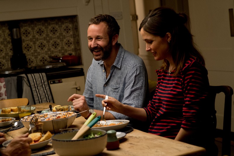 Mężczyzna i kobieta siedzą przy kuchennym stole, uśmiechają się i wspólnie jedzą posiłek. Różne naczynia i miski z jedzeniem są rozłożone przed nimi na drewnianym stole. Otoczenie wydaje się ciepłe i przytulne.