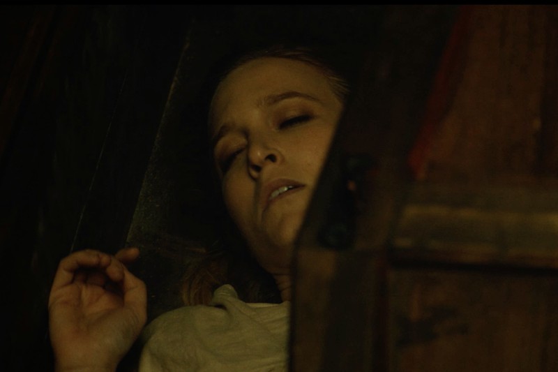 Kobieta z zamkniętymi oczami leży na plecach w słabym oświetleniu, częściowo zasłonięta drewnianą powierzchnią, co sugeruje, że może być nieprzytomna lub spać.