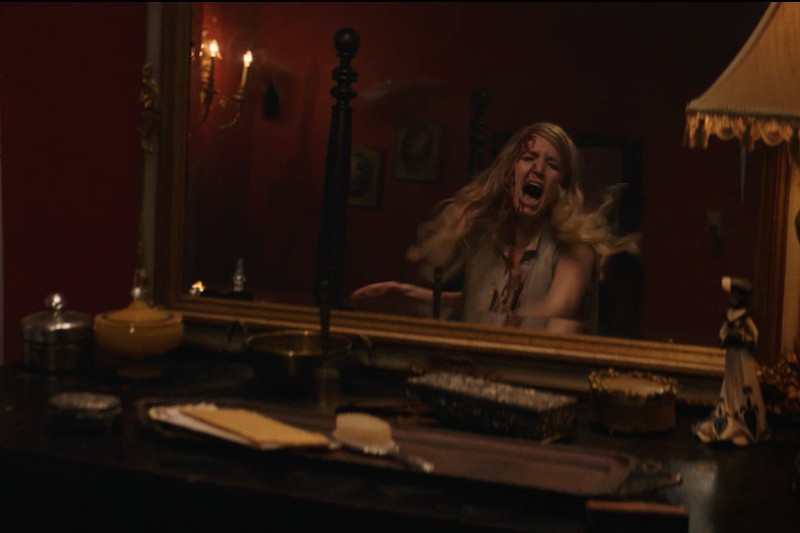 Kobieta o długich blond włosach i przerażonym wyrazie twarzy krzyczy przed lustrem w słabo oświetlonym pokoju z czerwonymi ścianami, w którym odbijają się różne przedmioty na komodzie.
