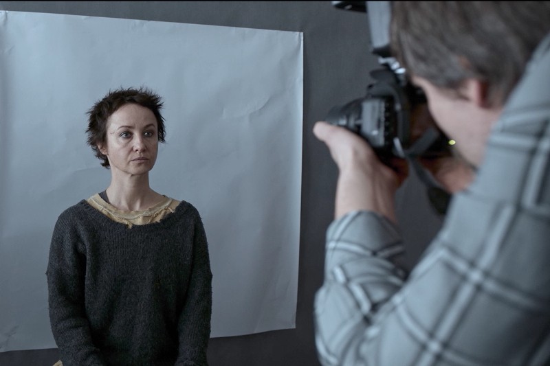 Kobieta z krótkimi włosami siedzi na tle pustego, białego tła, podczas gdy fotograf w kraciastej koszuli robi jej zdjęcie w studiu.