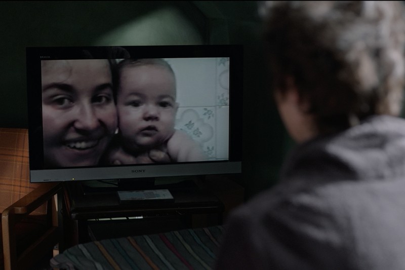 Osoba siedzi na łóżku i ogląda ekran telewizora wyświetlający zbliżenie uśmiechniętej kobiety trzymającej dziecko. Pokój jest słabo oświetlony.