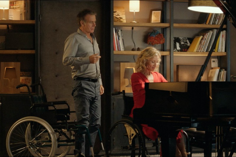 Mężczyzna stoi obok kobiety w czerwonej sukience, która siedzi na wózku inwalidzkim i gra na pianinie. W pobliżu znajduje się kolejny pusty wózek inwalidzki, a w tle półki z książkami i lampami.