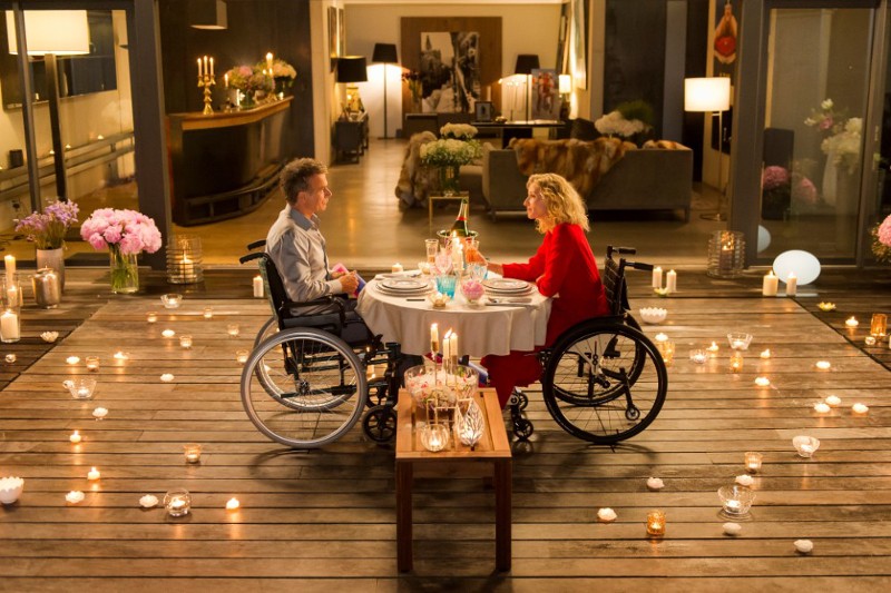 Mężczyzna i kobieta na wózkach inwalidzkich spożywają kolację przy świecach przy okrągłym stole na drewnianym tarasie, w otoczeniu kwiatów i licznych zapalonych świec. W tle widać przytulny, elegancki salon.
