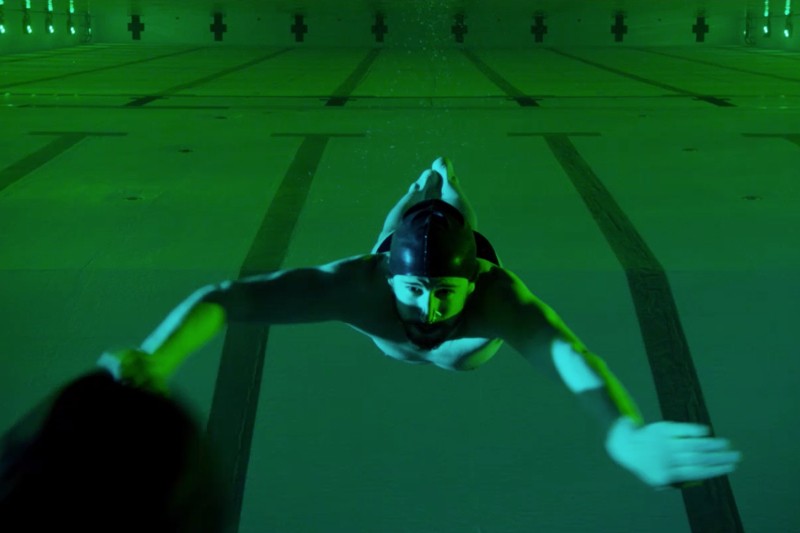 Pływak w czepku i goglach pływackich znajduje się pod wodą w basenie, wyciągając jedną rękę do przodu. Basen jest oświetlony zielonym światłem, a na dnie widoczne są oznaczenia torów.