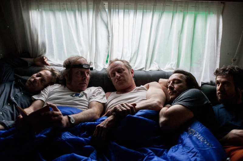 Pięciu mężczyzn leży blisko siebie pod niebieskim śpiworem na kanapie w słabo oświetlonym pokoju z białymi zasłonami. Wyglądają na zrelaksowanych i pochłoniętych rozmową.