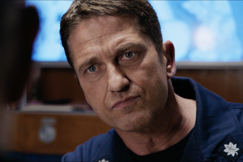 Mężczyzna z krótkimi brązowymi włosami i poważnym wyrazem twarzy uważnie przygląda się komuś poza kadrem. Ma na sobie ciemnoniebieski mundur z insygniami na kołnierzu. Tło jest lekko rozmyte.