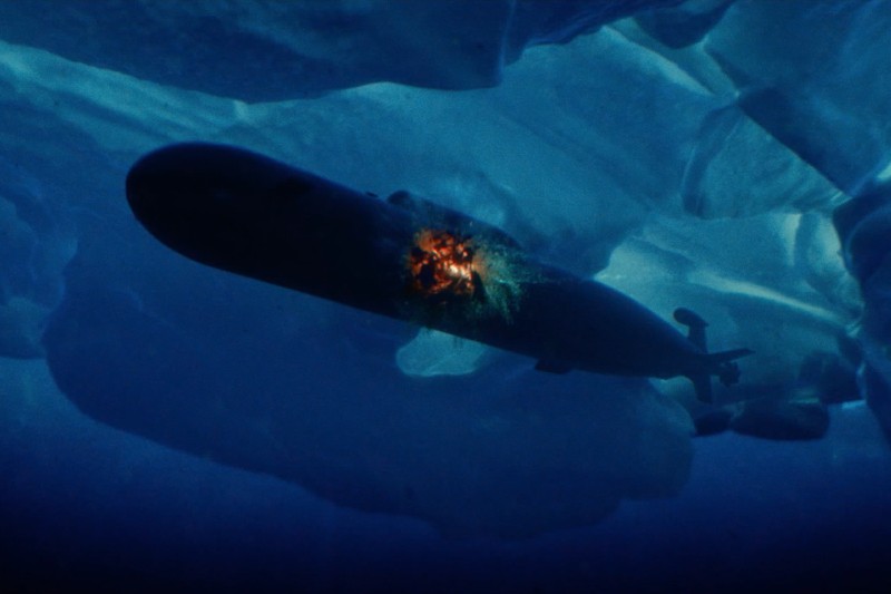Łódź podwodna pod wodą zostaje uderzona eksplozją w jej centrum, a wokół niej znajdują się lodowe formacje. Pomarańczowy wybuch kontrastuje z ciemnoniebieskimi tonami lodu i wody.