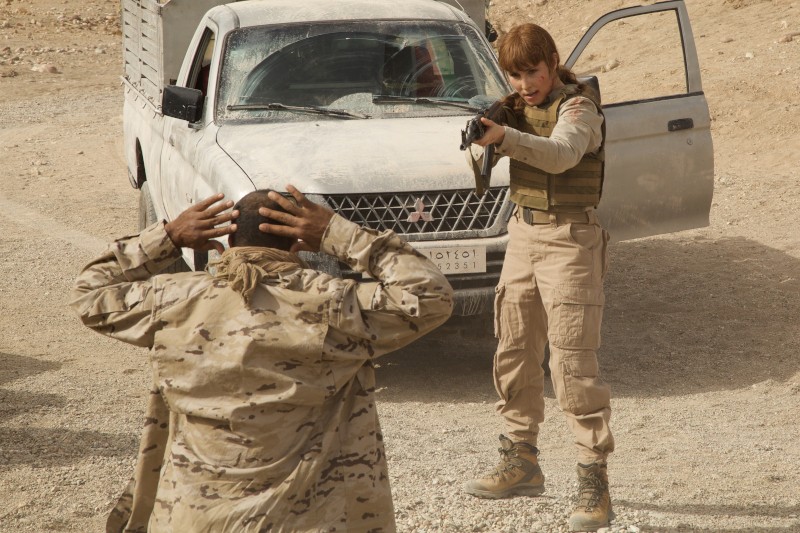 Osoba w stroju wojskowym celuje z broni krótkiej w inną osobę, która trzyma ręce na głowie i stoi obok białej, zakurzonej ciężarówki z otwartymi drzwiami w otoczeniu przypominającym pustynię.