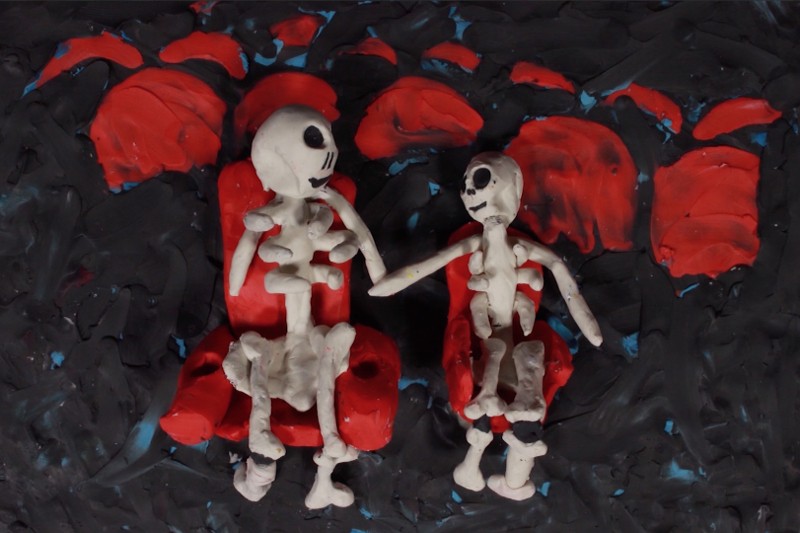 Dwie szkieletowe figury wykonane z gliny siedzą na czerwonych krzesłach na ciemnym, teksturowanym tle z czerwonymi i niebieskimi akcentami. Jeden szkielet wydaje się większy, prawdopodobnie dorosły, a drugi jest mniejszy, przypomina dziecko.