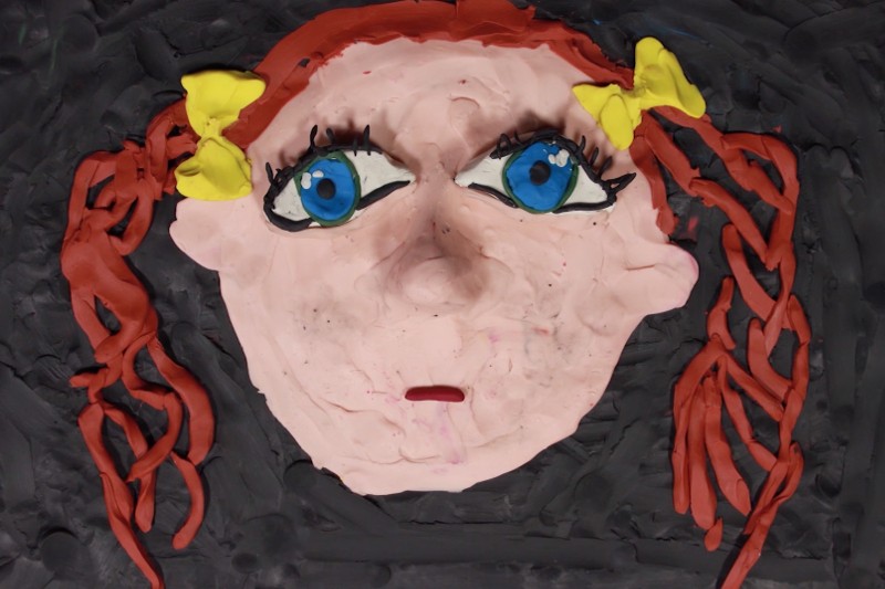 Twarz wykonana z gliny, duże niebieskie oczy, czerwone warkocze związane żółtymi kokardkami i neutralny wyraz twarzy na czarnym tle.