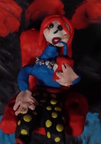 Gliniana figurka z długimi rudymi włosami, niebieską koszulą i czarnymi spodniami w żółte kropki siedzi na czerwonym krześle, trzymając czerwony kubek ze słomką. Tło jest ciemne z czerwonymi akcentami.