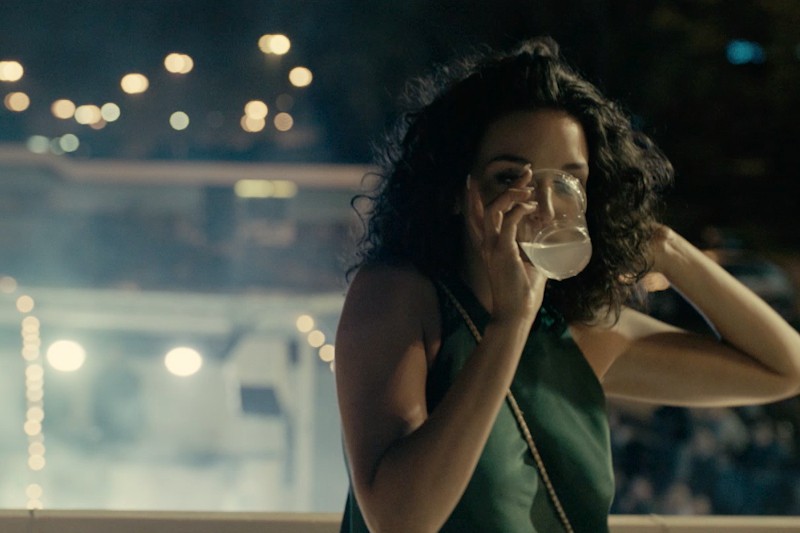 Kobieta o kręconych, ciemnych włosach, ubrana w zielony top bez rękawów, pije z przezroczystego szkła nocą, siedząc na balkonie, na tle rozmazanych świateł miasta.