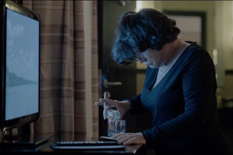 Kobieta w czarnej sukience nalewa drinka do szklanki w pobliżu telewizora pokazującego scenę z natury. Stoi w słabo oświetlonym pokoju, patrzy w dół, z zapalonym papierosem w dłoni.