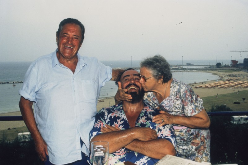 Trzy osoby stoją na balkonie przy plaży; starsza kobieta całuje brodatego mężczyznę w policzek i ściska jego twarz, podczas gdy starszy mężczyzna uśmiecha się obok nich. Nastrój jest radosny i serdeczny, w tle widać morze.