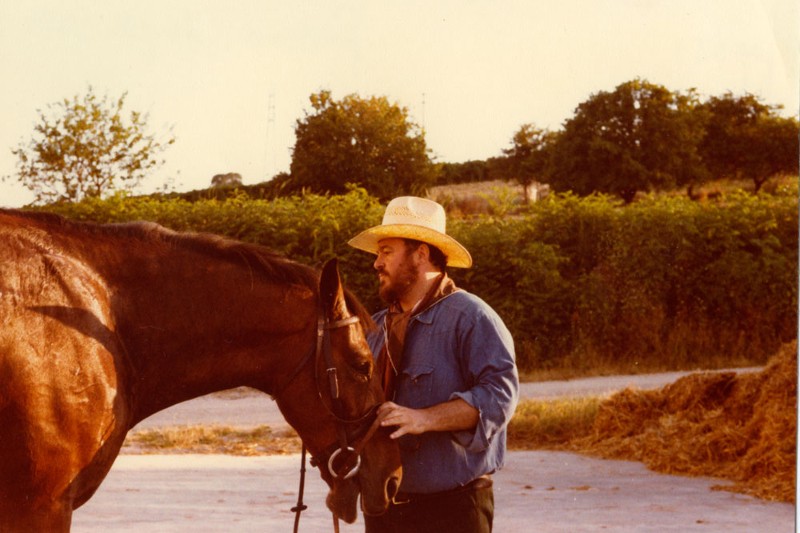 Brodaty mężczyzna w słomkowym kapeluszu i niebieskiej koszuli stoi na zewnątrz, delikatnie trzymając uzdę brązowego konia. W tle w słoneczny dzień widać drzewa i krzewy.