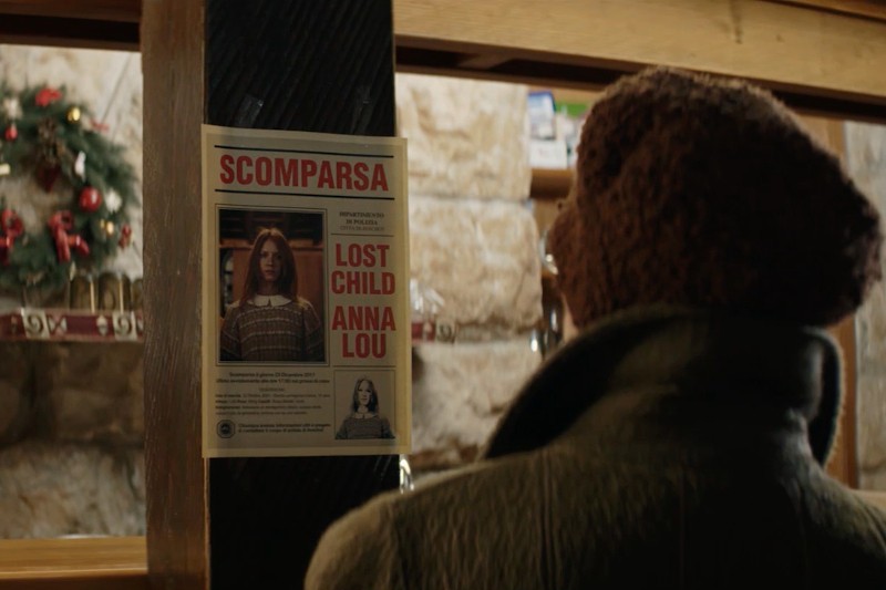 Osoba w brązowym płaszczu i kapeluszu patrzy na plakat zaginionego dziecka na ścianie. Na plakacie widnieje napis Scomparsa i Zaginione Dziecko Anna Lou, a w pobliżu widać zdjęcie młodej dziewczynki i świąteczne dekoracje.