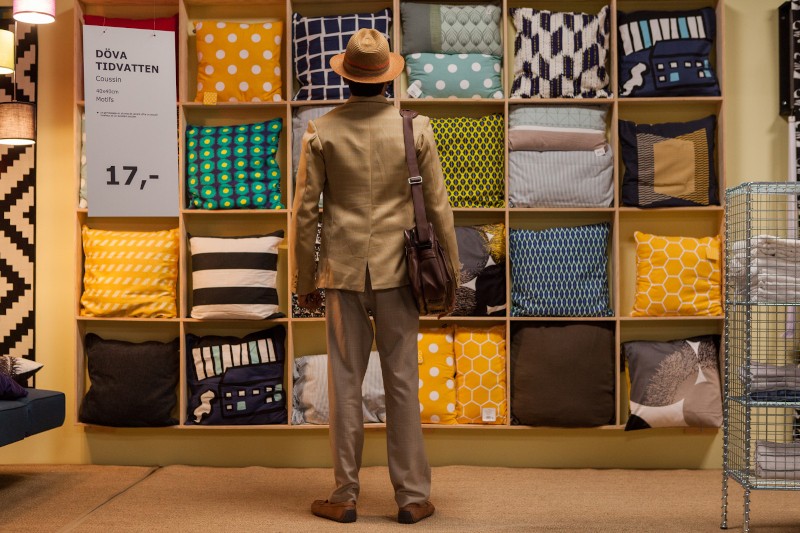 Osoba w beżowym garniturze i kapeluszu stoi naprzeciwko ekspozycji kolorowych, wzorzystych poduszek ułożonych na półkach w sklepie. Osoba ma brązową torbę na ramię i jest odwrócona tyłem do kamery.