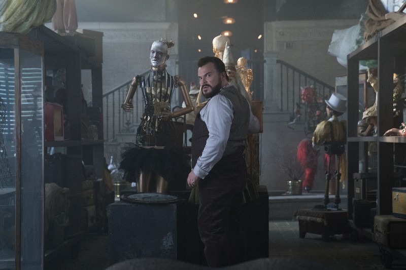 Mężczyzna w kamizelce i fartuchu stoi w słabo oświetlonym pomieszczeniu wypełnionym manekinami, kostiumami i różnymi osobliwościami, patrząc przez ramię z poważnym wyrazem twarzy. W tle widać schody i więcej przedmiotów.