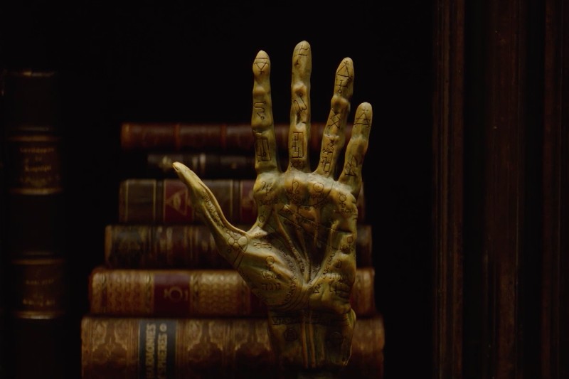 Mosiężna rzeźba przedstawiająca ludzką dłoń z wygrawerowanymi liniami i symbolami chiromancji jest wystawiona pionowo przed stosem starych, oprawionych w skórę książek na ciemnym tle.