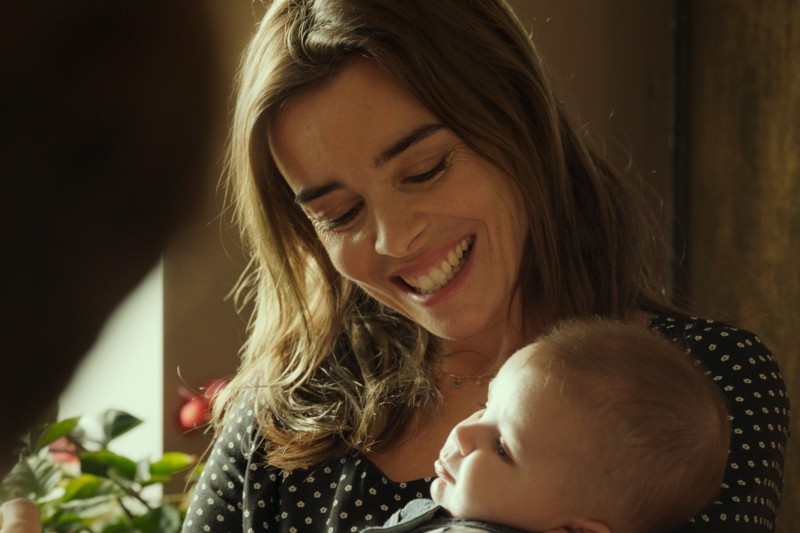 Kobieta z długimi brązowymi włosami, ubrana w czarny top w białe kropki, uśmiecha się ciepło, trzymając w ramionach dziecko. Dziecko patrzy na nią. Z boku pada na nie miękkie światło.