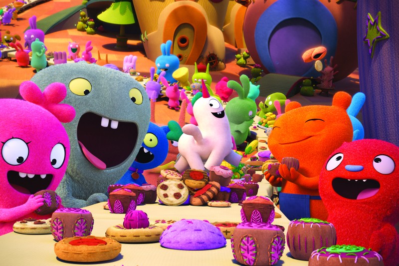 Kolorowe, animowane postacie potworów z szerokimi uśmiechami siedzą przy stole pełnym ciast i deserów, ciesząc się ożywioną ucztą na świeżym powietrzu w tętniącym życiem, fantastycznym otoczeniu z jeszcze większą liczbą stworzeń w tle.