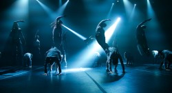 Tancerze wykonują dramatyczne skoki i wygięcia na słabo oświetlonej scenie, przy czym niebieskie światło i reflektory tworzą dynamiczne cienie i napiętą atmosferę.