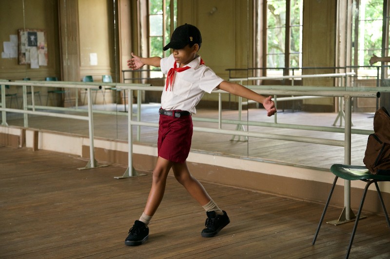 Dziecko ubrane w czarną czapkę, czerwony szalik, białą koszulkę i czerwone spodenki ćwiczy pozę taneczną z wyciągniętymi ramionami w studiu z drewnianą podłogą i lustrami ściennymi.