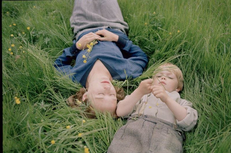 Kobieta i dziecko leżą w wysokiej zielonej trawie, twarzą do góry. Kobieta trzyma żółte kwiaty, a dziecko bada źdźbło trawy, oboje wydają się zrelaksowani i spokojni.