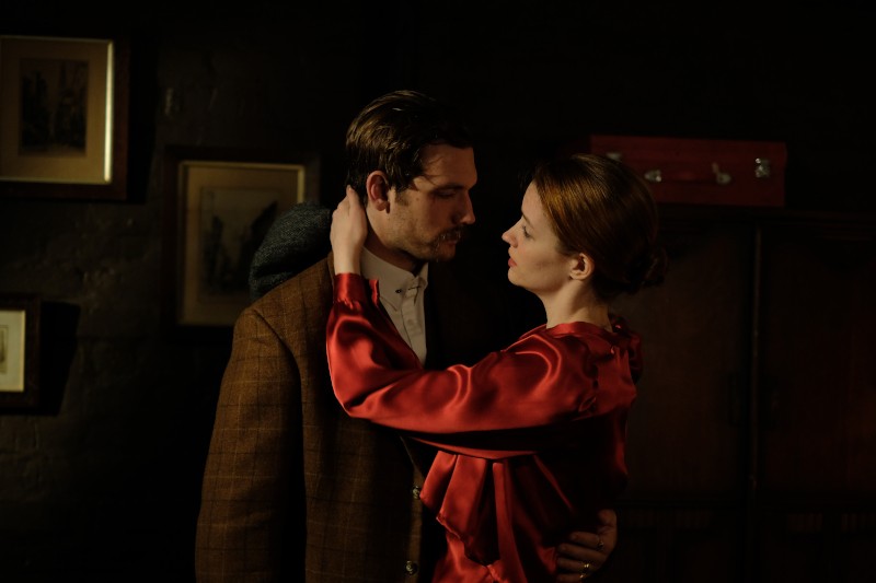 Mężczyzna i kobieta obejmują się blisko w słabo oświetlonym pokoju. Kobieta ubrana w czerwoną satynową sukienkę patrzy na mężczyznę ubranego w brązową kraciastą marynarkę i białą koszulę. W tle widać oprawione zdjęcia i ciemne meble.