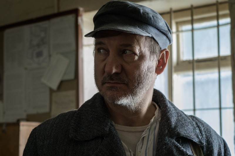 Mężczyzna w średnim wieku z brodą, ubrany w szarą czapkę i płaszcz, stoi w pomieszczeniu przy zakratowanym oknie, patrząc w bok z poważnym wyrazem twarzy. Za nim na tablicy ogłoszeń przypięto papiery.