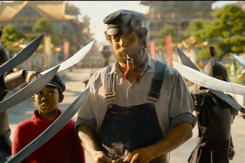 Mężczyzna w kombinezonie i czapce, palący fajkę, stoi pewnie otoczony wymierzonymi w niego mieczami. Obok niego stoi młody chłopiec w czerwonym swetrze, patrząc w górę, z budynkami i zielenią w tle.