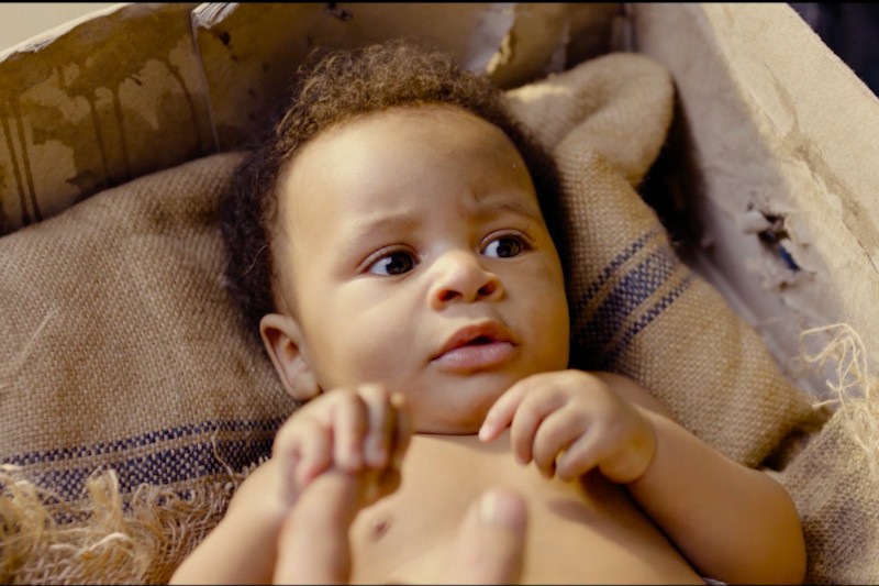 Dziecko o kręconych włosach leży na płótnie jutowym wewnątrz tekturowego pudełka i patrzy w górę z lekko zaniepokojonym wyrazem twarzy.