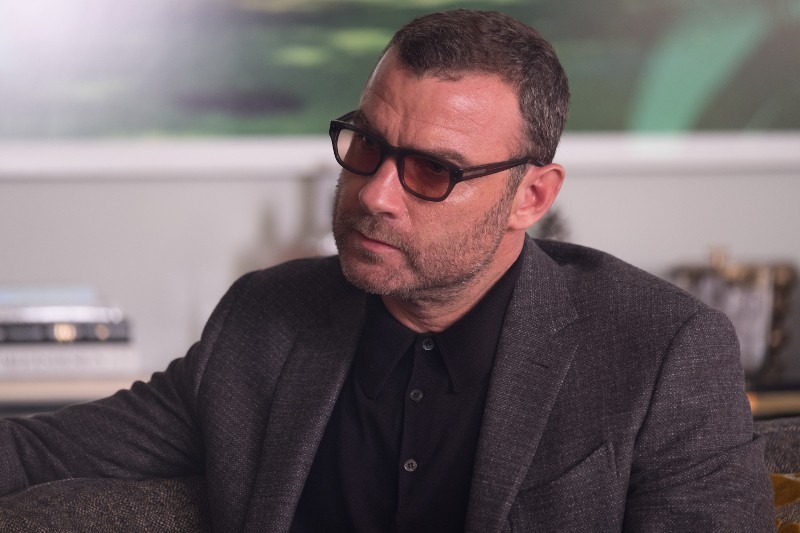 Mężczyzna z krótkimi włosami i brodą, ubrany w ciemne okulary, ciemną koszulę z kołnierzykiem i szary blezer, siedzi wewnątrz, patrząc w bok z poważnym wyrazem twarzy.