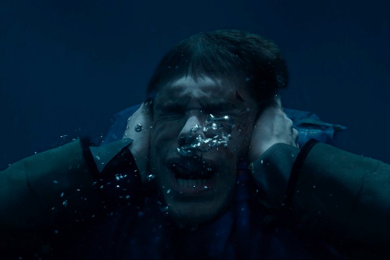 Mężczyzna pod wodą, z zamkniętymi oczami i otwartymi ustami, jakby krzyczał, z rękami mocno przyciśniętymi do obu stron głowy. Z jego ust wydobywają się bąbelki, a jego wyraz twarzy wydaje się być zmartwiony.