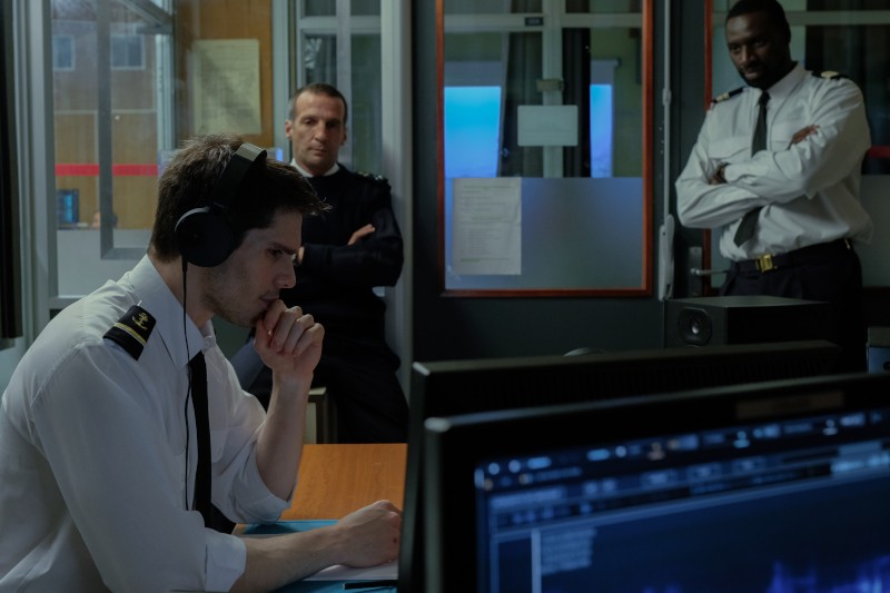 W pokoju kontrolnym znajduje się trzech umundurowanych mężczyzn; jeden z nich siedzi ze słuchawkami na uszach i wydaje się wpatrywać w ekran komputera, podczas gdy pozostali dwaj stoją za nim i poważnie obserwują.