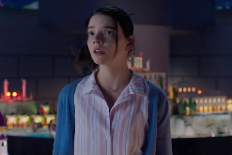 Młoda kobieta o ciemnych włosach, ubrana w pasiastą koszulę i niebieski kardigan, stoi na tle rozmytego tła, na którym widać miniaturowe budynki i światła. Patrzy w górę z zaniepokojonym wyrazem twarzy.