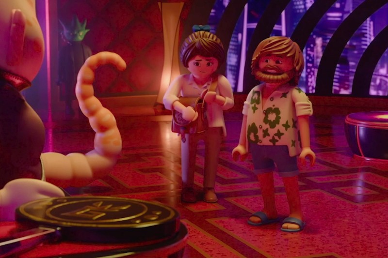 Dwie figurki Playmobil, jedna z brodą, ubrana w kwiecistą koszulę i spodenki, a druga z kucykiem i białą koszulą, stoją w kolorowym, futurystycznym pokoju z odważnym oświetleniem i wzorzystą podłogą.