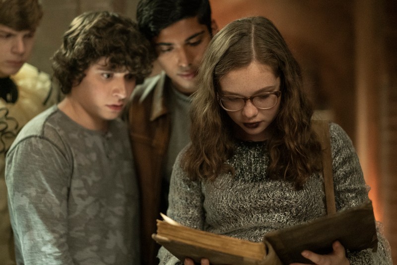 Czwórka nastolatków stoi blisko siebie w słabo oświetlonym pokoju, uważnie wpatrując się w starą, otwartą książkę, którą trzyma dziewczyna w okularach i z kręconymi włosami. Nastrój wydaje się napięty i tajemniczy.