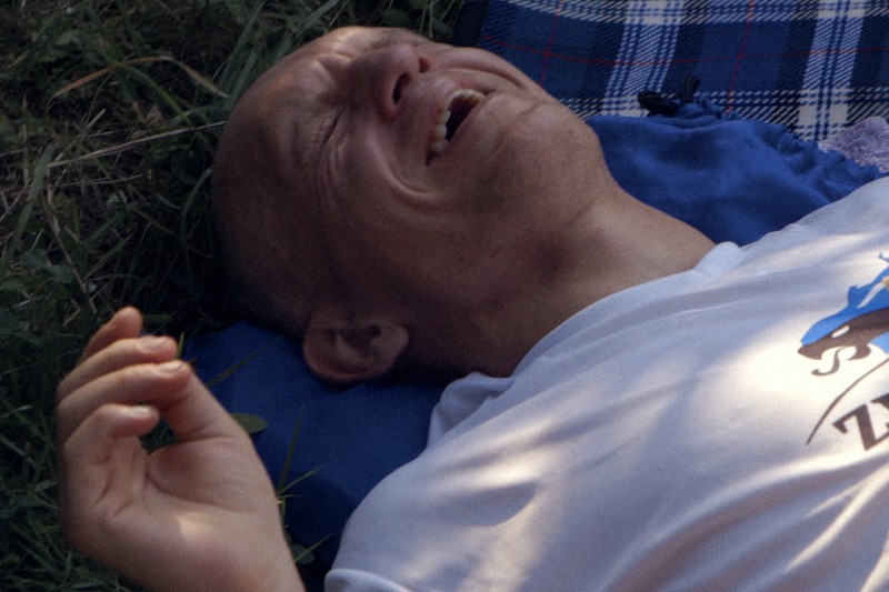 Osoba z ogoloną głową leży na niebieskim kocu na zewnątrz, uśmiechnięta lub śmiejąca się z zamkniętymi oczami, ubrana w białą koszulkę. Wokół niej widać trawę.