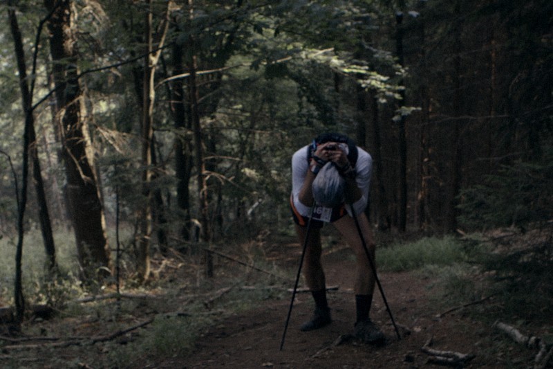 Osoba z kijkami trekkingowymi pochyla się wyczerpana na leśnej ścieżce, mając na sobie numer startowy i sprzęt sportowy. Światło słoneczne przesącza się przez drzewa, tworząc nastrojową, zacienioną atmosferę w lesie.