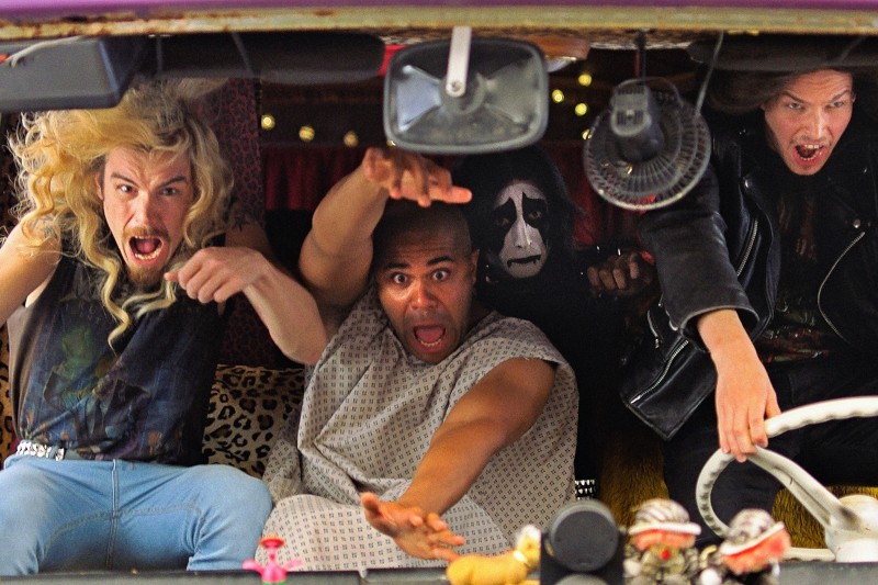 Trzech mężczyzn o dzikich minach siedzi w chaotycznym samochodzie; jeden nosi szpitalny fartuch, drugi ma długie blond włosy, a trzeci ma pomalowaną twarz. Kierowca w skórzanej kurtce gorączkowo kieruje. Przedmioty zagracają deskę rozdzielczą.