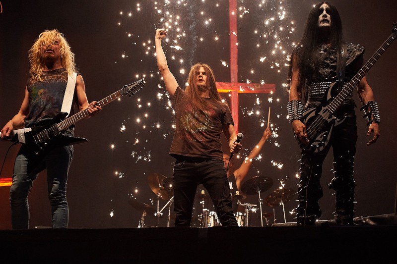 Trzech muzyków metalowych występuje na scenie z gitarami elektrycznymi; wokalista podnosi pięść, gdy za nimi wybuchają iskry. Noszą ciemne, odważne stroje i mocny makijaż, a w tle widać perkusistę.