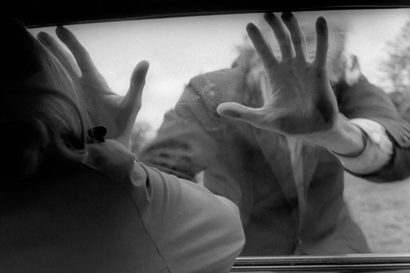 Czarno-biały obraz przedstawia osobę siedzącą w samochodzie, przyciskającą ręce do szyby, podczas gdy inna osoba na zewnątrz wyciąga w jej stronę ręce, co wywołuje poczucie napięcia lub strachu.