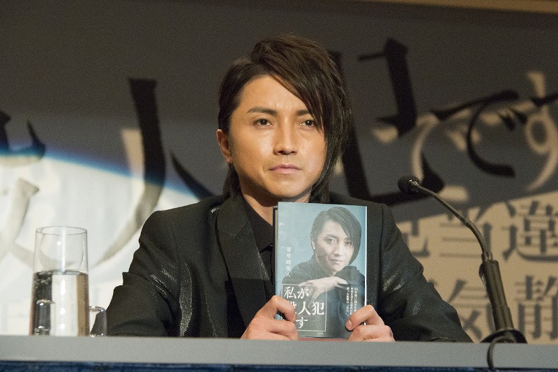 Osoba o ciemnych włosach ubrana w czarny garnitur siedzi przy stole, trzymając książkę z własnym portretem na okładce. Na stole stoi szklanka wody i mikrofon, a w tle rozmazany baner.