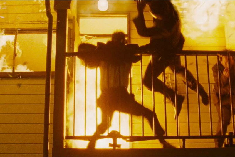 Dwie osoby wyskakują z balkonu, gdy płomienie ogarniają budynek za nimi, co sugeruje dramatyczną ucieczkę z pożaru. Scena jest jasno oświetlona przez płomienie.